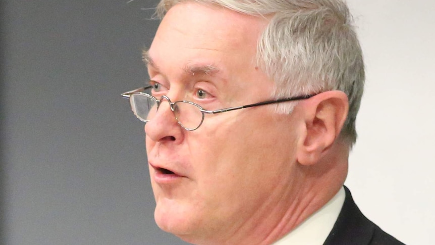 Robert Cribb wearing suit, blue time and silver rim glasses stands with grey wall in background.