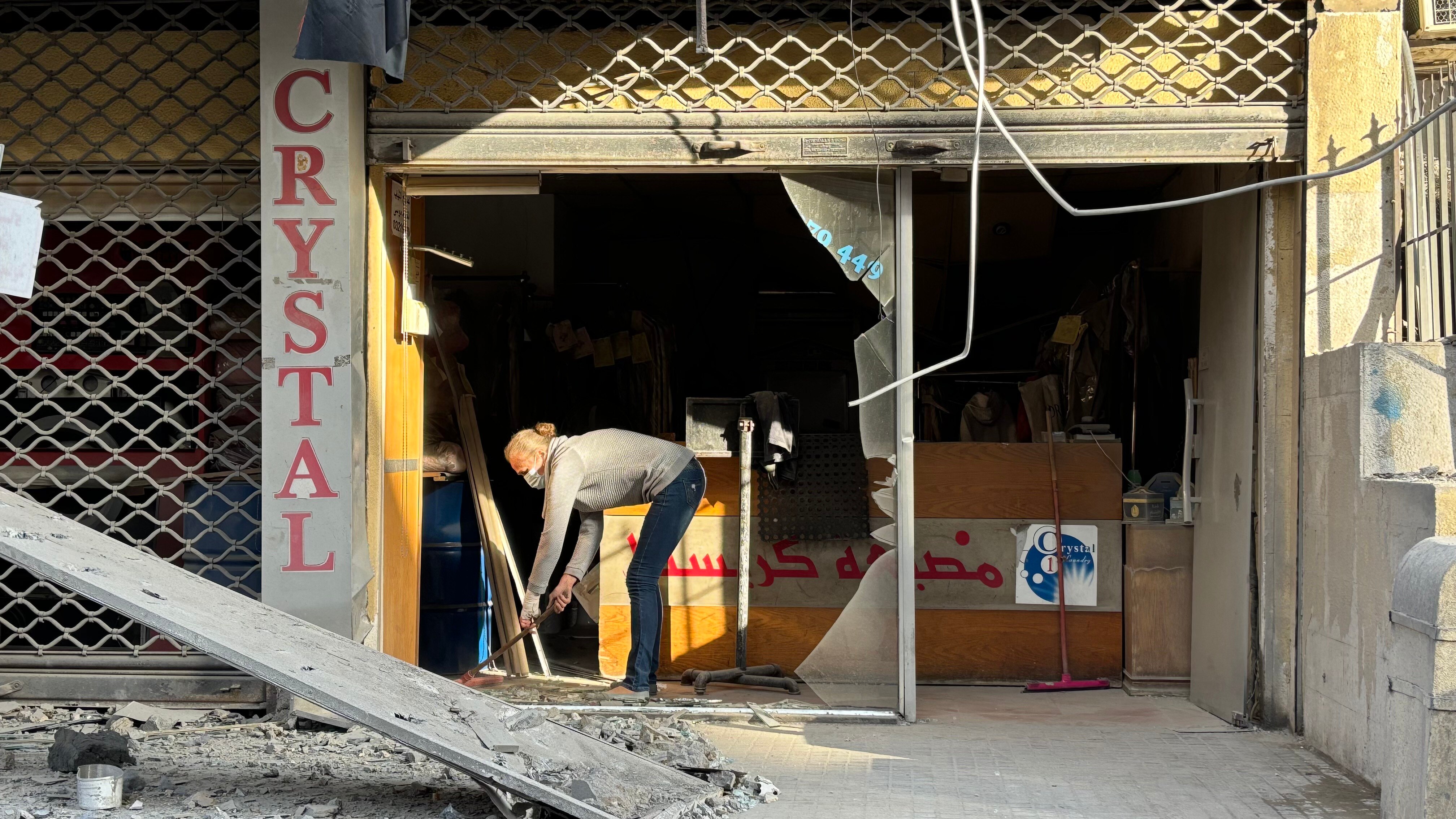 A photo of a slim woman sweeping her damaged shop with a broom.