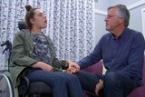 Ashleigh sit in a wheelchair and holds her father's hand. He is sitting near her on a sofa, looking at her.