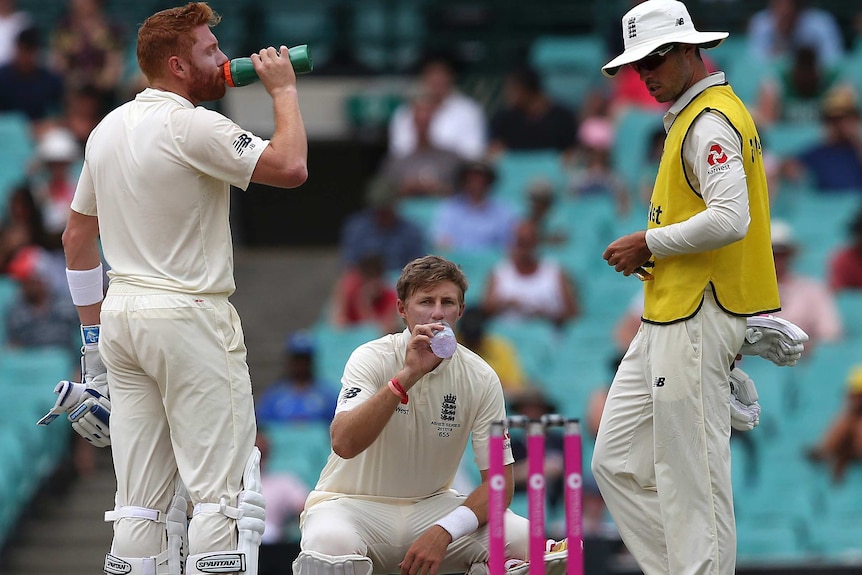 Joe Root squats while drinking water out on the field, flanked by two other players.