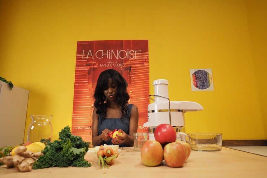 Young Black woman sits at table in bright yellow room cutting fruit, with poster of Godard's film La Chinoise behind her.