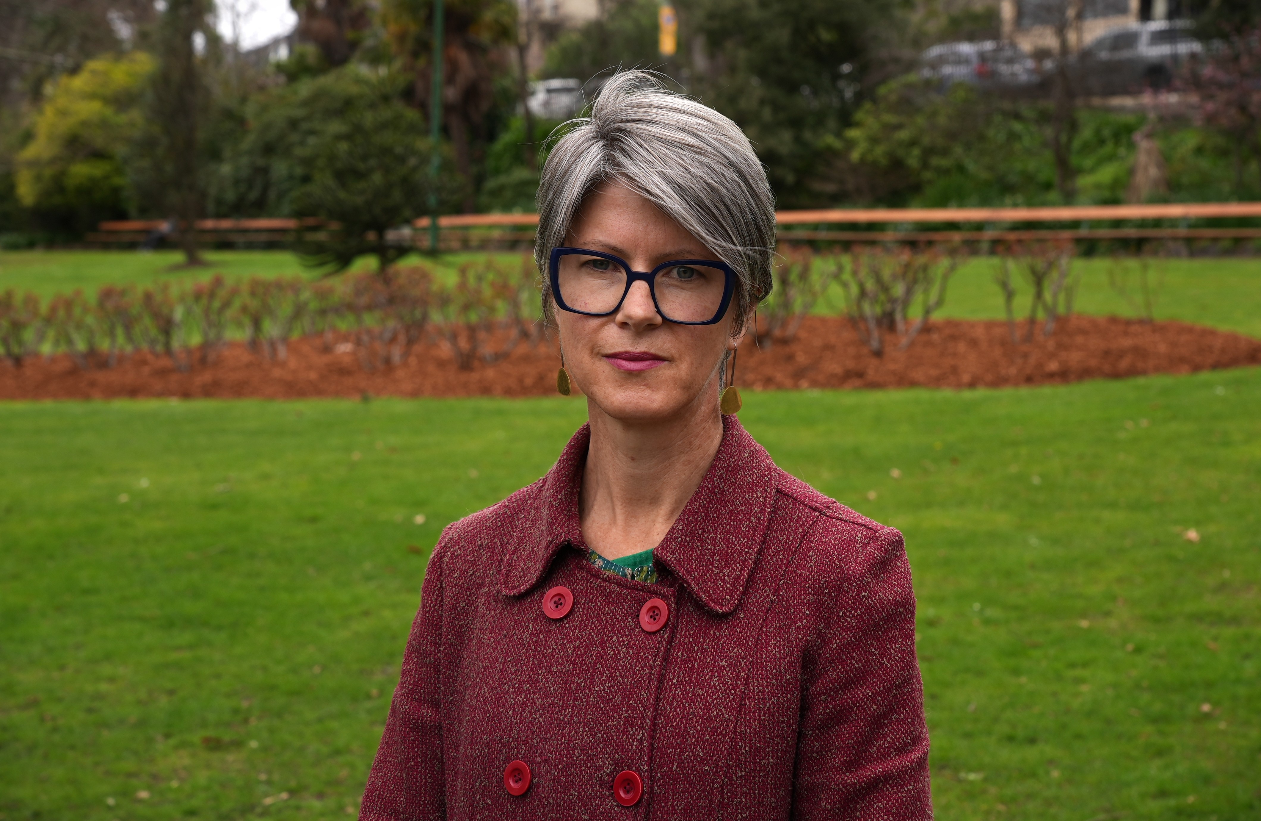 Cecily Rosol se encuentra en un parque con un abrigo rojo oscuro, gafas gruesas y aretes colgantes.