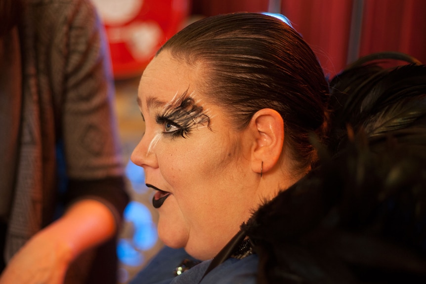 A photo taken from side on of a woman with elaborate stage makeup, including black lipstick, long eyelashes and eye makeup.
