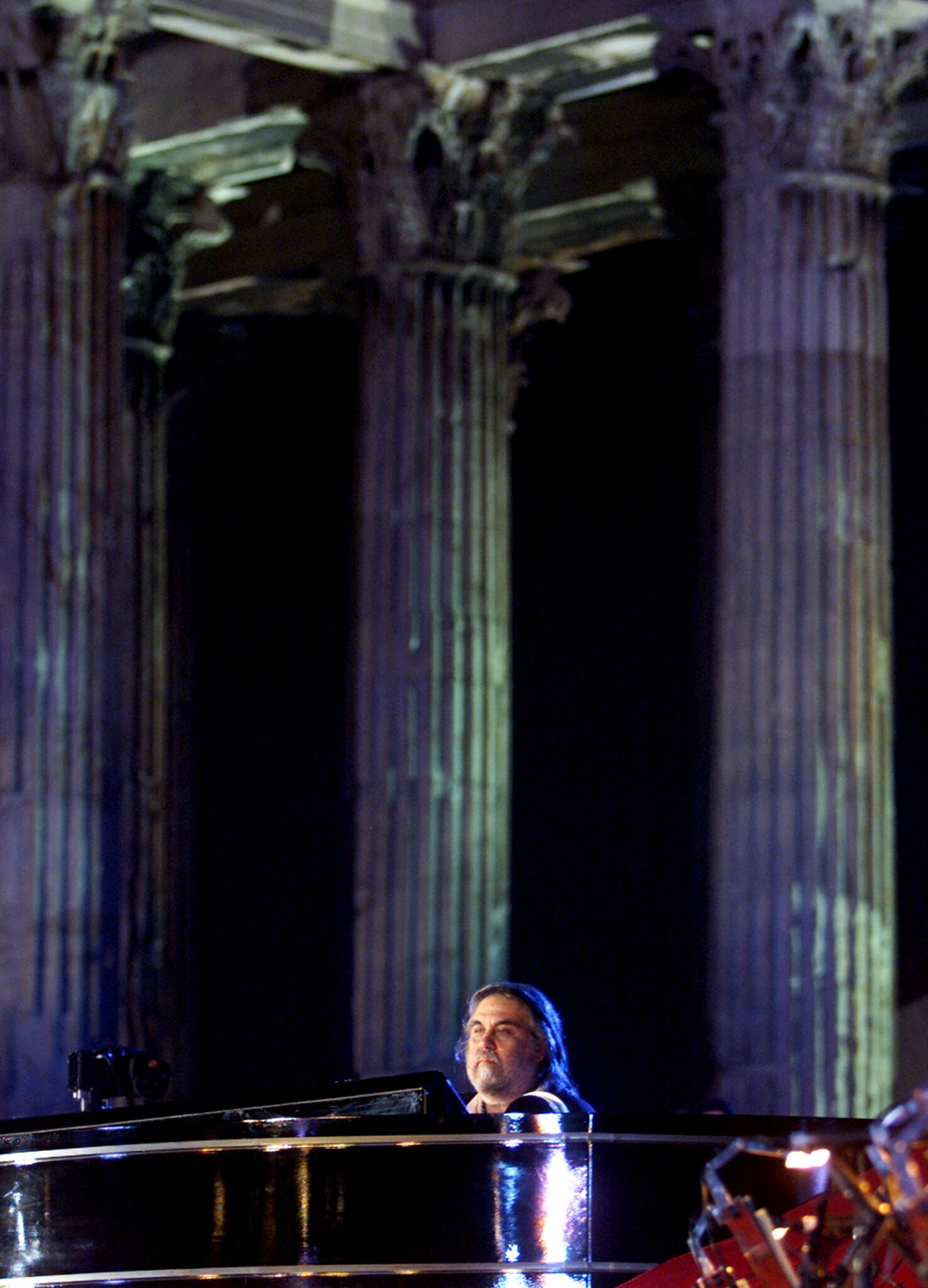 A man sits at a piano with a backdrop of three Greek columns behind him