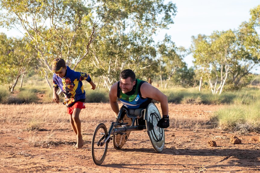White man in 3-wheel racing wheelchair and young Aboriginal boy poised to race in red dirt desert landscape.