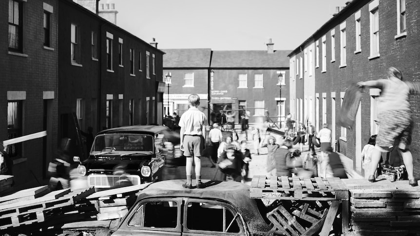 A black and white image of a boy in 60s-style clothing standing on top of a car at the end of a barricaded street