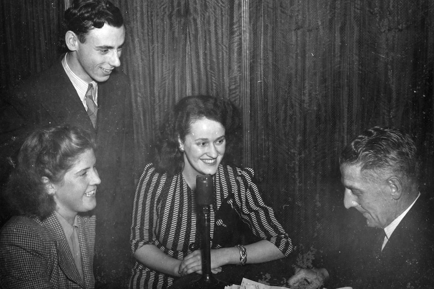 A black and white photo of three young people facing an older man sitting at a table. The young people are smiling.
