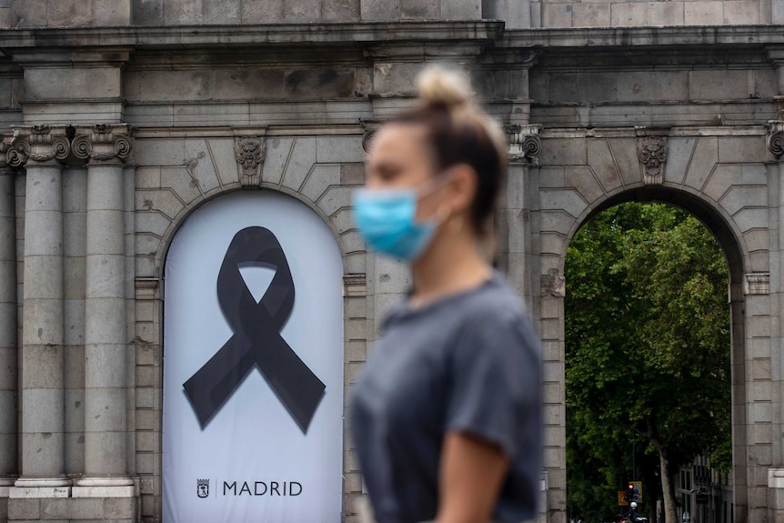 a woman in a mask walks in front of a poster displaying a black ribbon.