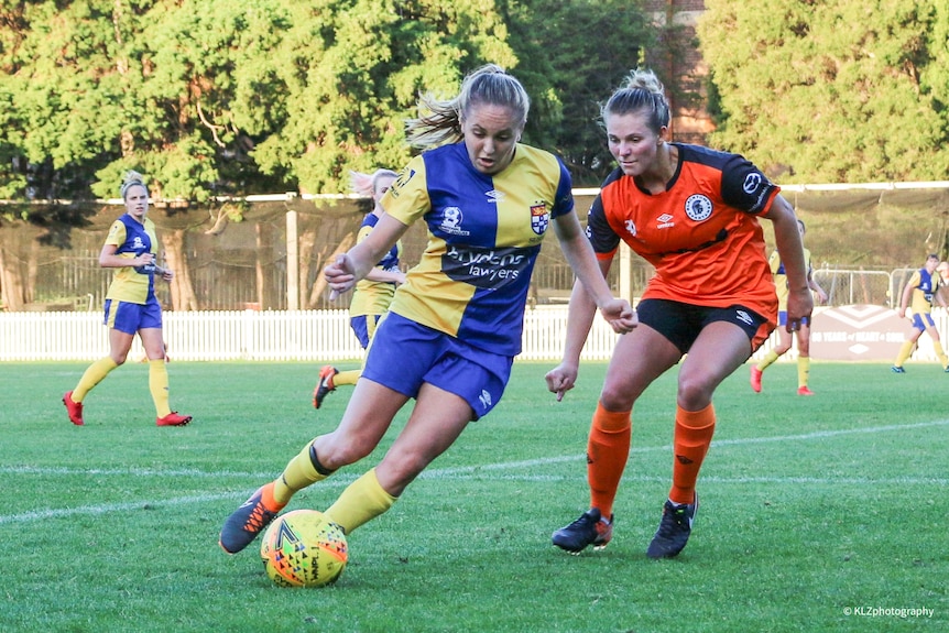 A female soccer player wearing blue and yellow looks at the ball near another player in orange and black