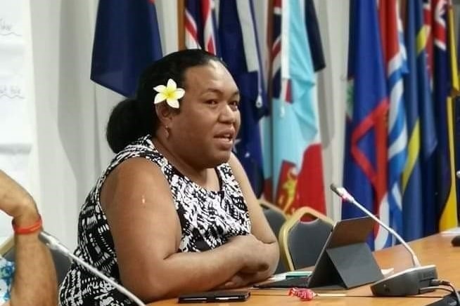 A Fijian woman sits at a desk speaking into a microphone with flags draped in background