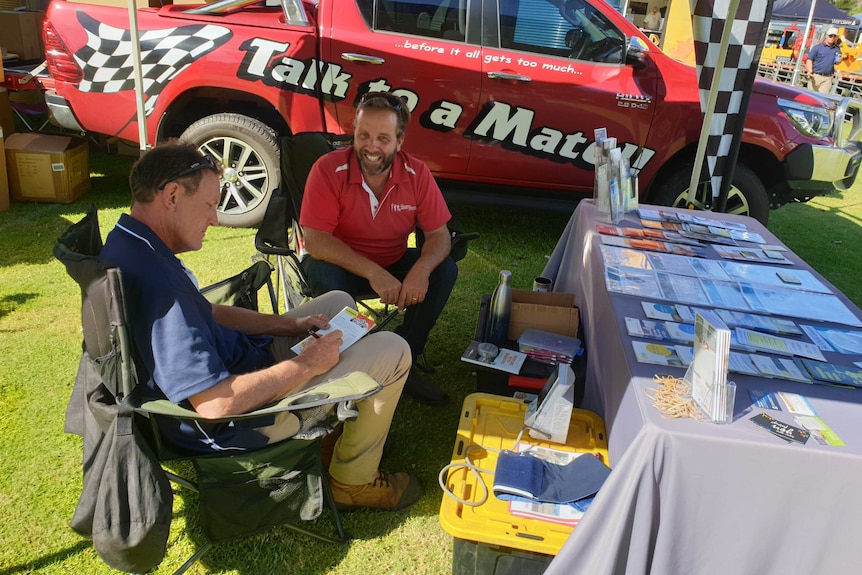 Two men sitting by a ute with a table full of flyers.