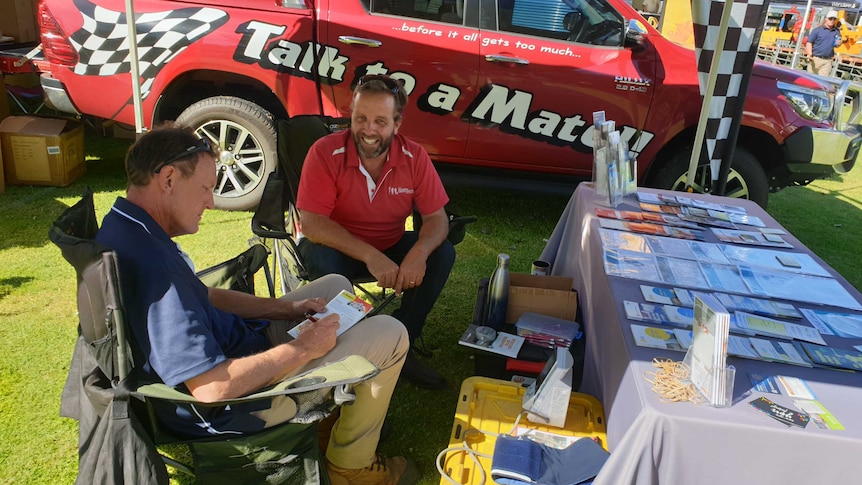 Two men sitting by a ute with a table full of flyers.