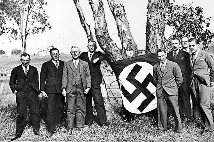 Eight men pose for a photo beneath a tree with a swastika flag wrapped around it