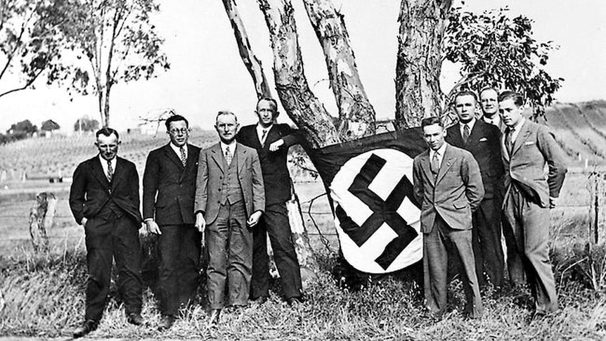 Eight men pose for a photo beneath a tree with a swastika flag wrapped around it