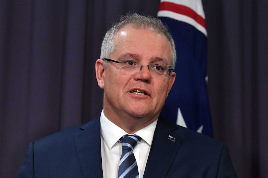 Scott Morrison looks to the right as he stands in front of a blue background mid-speech. An Australian flag is also behind him.