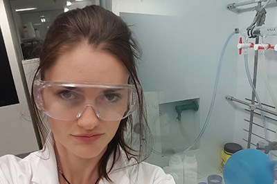 A young woman wearing a white lab coat and clear plastic safety glasses, in a laboratory.