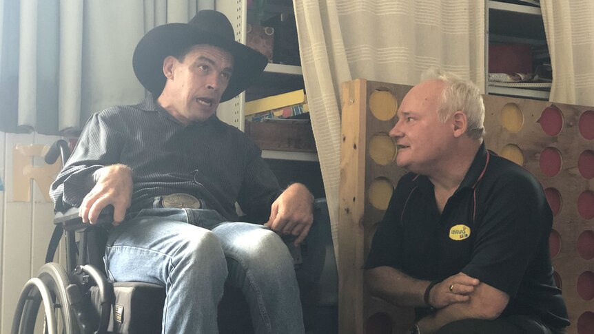 A man in an Akubra-style hat, sitting in a wheelchair, speaks to a man kneeling beside him.