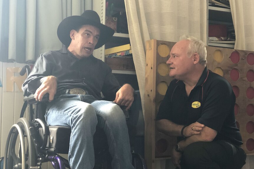 A man in an Akubra-style hat, sitting in a wheelchair, speaks to a man kneeling beside him.