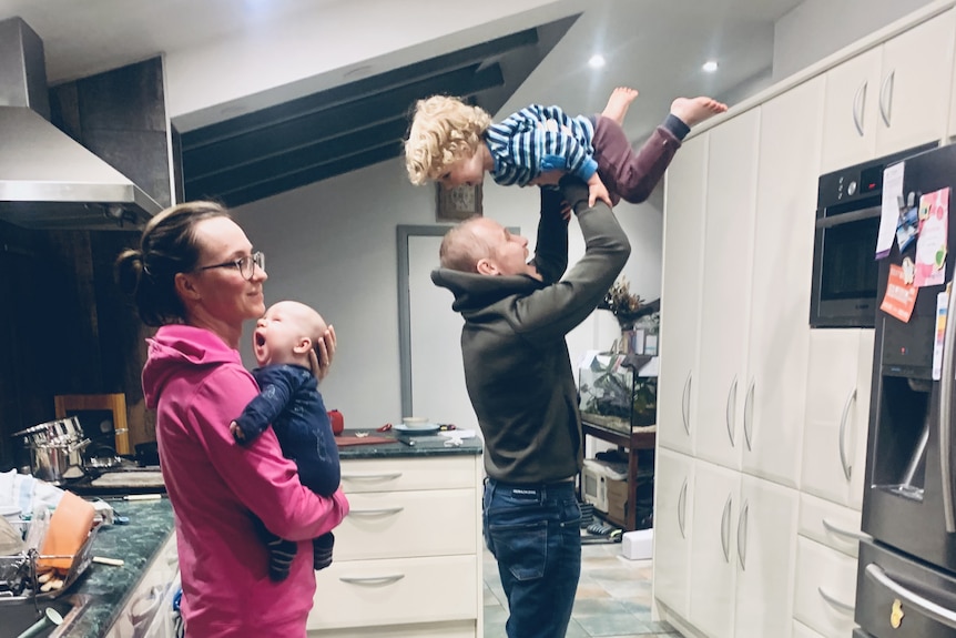 Michael picks up Bryn over his head while Kiri holds their baby, all standing in the kitchen.
