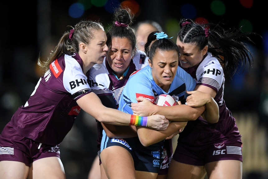 A rugby league player stands crouched over while being tackled by three opponents in the Women's State of Origin match.