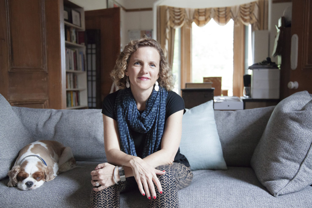 Sally Rippen, with short curly hair, black shirt and brown pants, sits on a couch smiling slightly. A dog sits next to her.