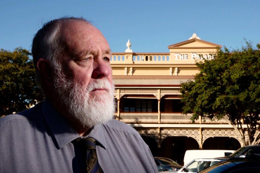 Senior man stand in foreground and looks off into the stance. Heritage hotel with federation era verandah is seen in background