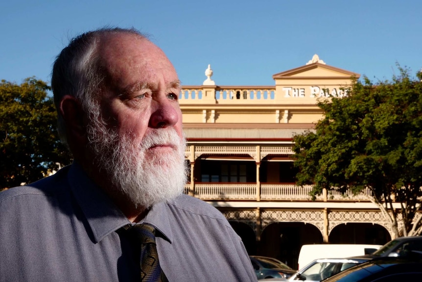 Senior man stand in foreground and looks off into the stance. Heritage hotel with federation era verandah is seen in background