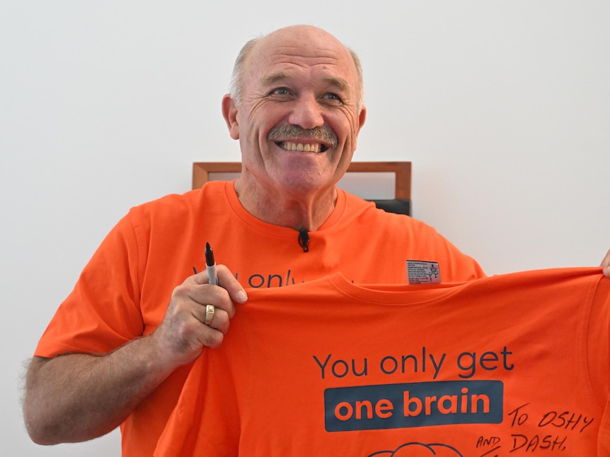 Former rugby league great Wally Lewis smiles for the cameras as he holds an orange t-shirt tagged "you only get one brain". 