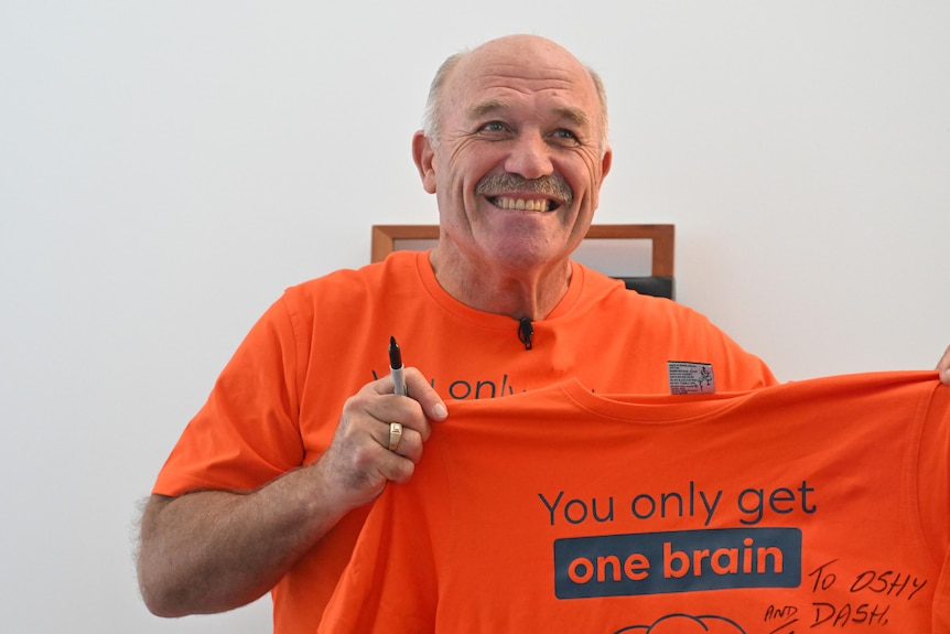 Former rugby league great Wally Lewis smiles for the cameras as he holds an orange t-shirt tagged "you only get one brain". 