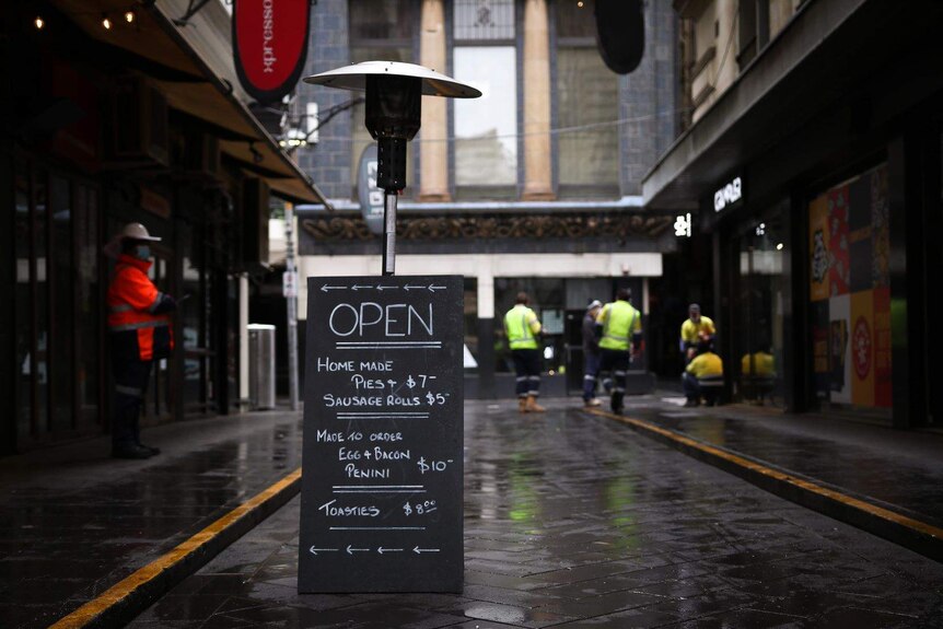 A wet and dark Melbourne laneway, with a cafe sign saying it is open for take-away, and workers standing behind eating.