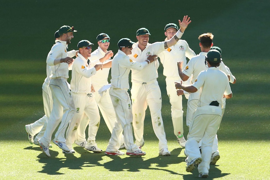 Cricketers, wearing cricket whites and baggy green caps, huddle and high-five after winning the test match