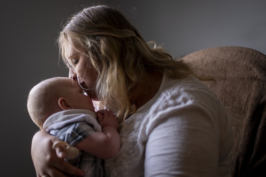 A mother kisses the forehead of her baby in soft window light.