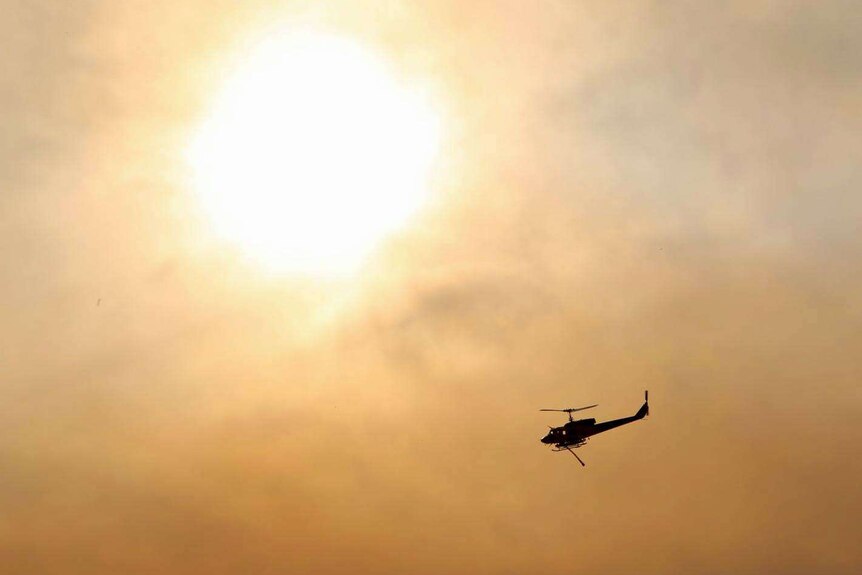 A helicopter water bomber flies near the sun in a smoke-filled sky.