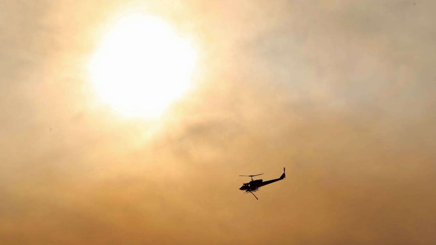 A helicopter water bomber flies near the sun in a smoke-filled sky.
