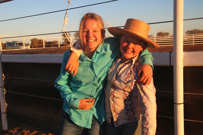 Two young girls stand with their arms around each other in front of a rodeo arena