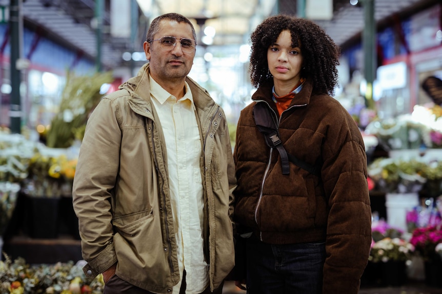 Robert, left, wears a beige jacket, cream shirt and glasses and stands in a market aisle next to Elsie, who wears a brown coat.