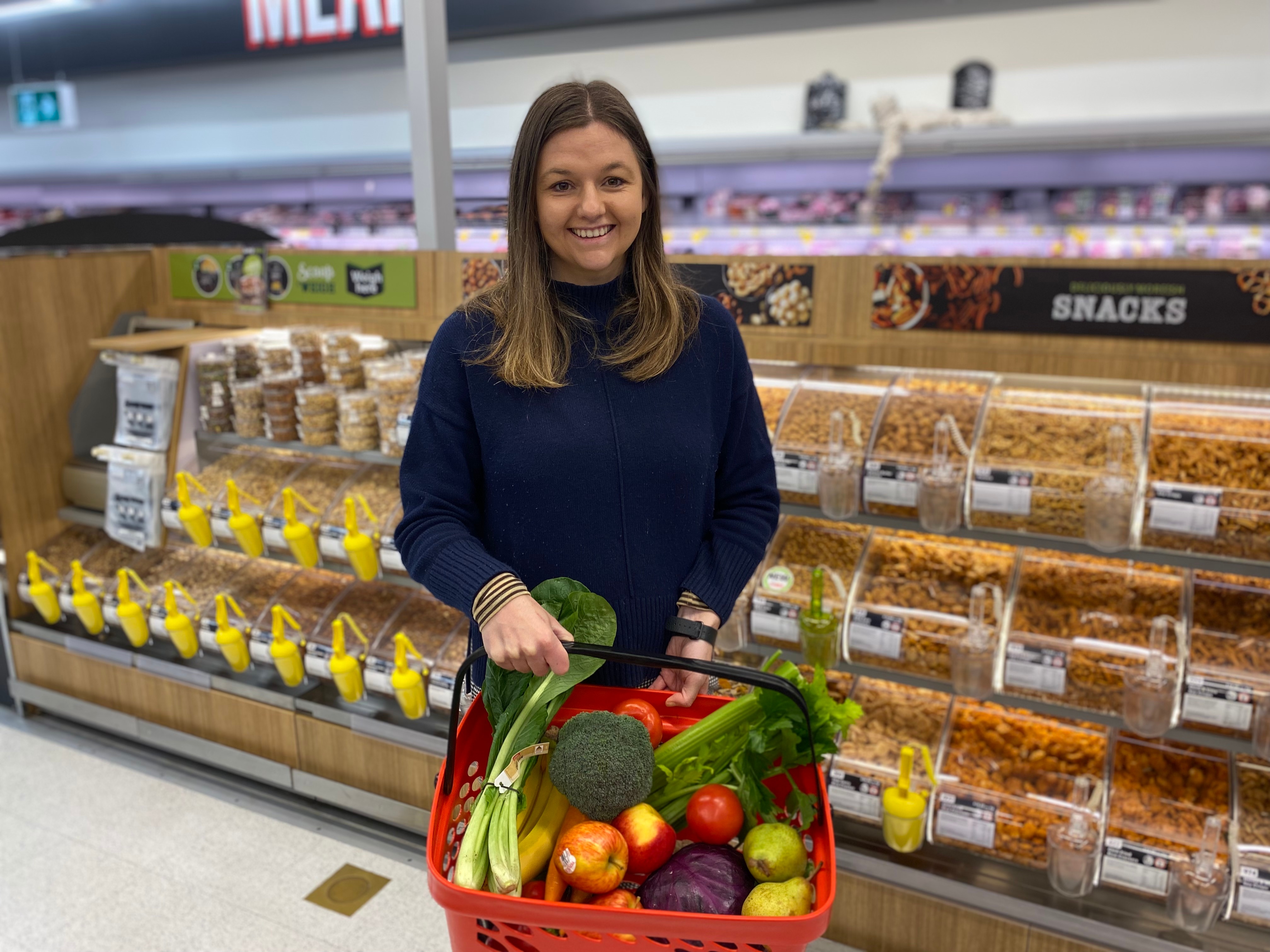A young woman holds a basket of vegetables in front of nut displays at Coles