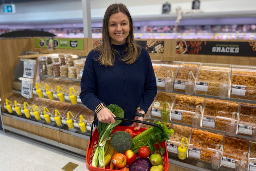 A young woman holds a basket of vegetables in front of nut displays at Coles