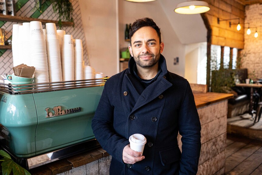 A man in a blue peacoat holds a takeaway coffee cup next to a barista machine
