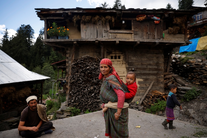 A mother, with her child strapped to her back, stands in front of her house, with her husband sitting to the left.