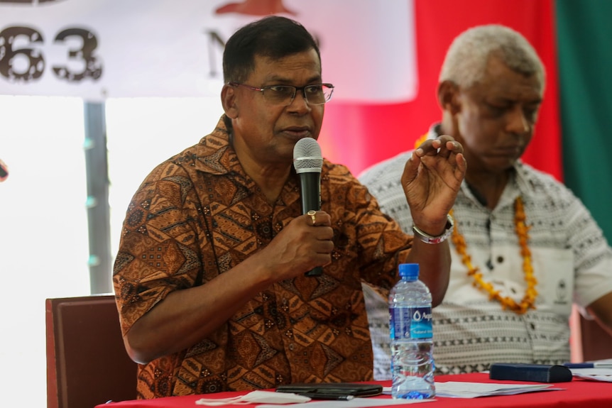 A man wearing a brown bula shirt speaks into a microphone sitting at a table with another person