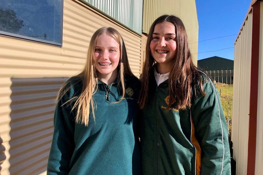 Two female high school students dressed in forest green jumpers smile at the camera. They stand near a corrugated iron shed.