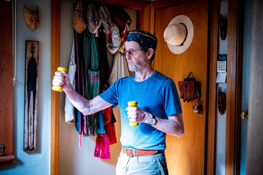 Karl Schurr stands by a door with hats and scarves hanging off it, while using dumbells.