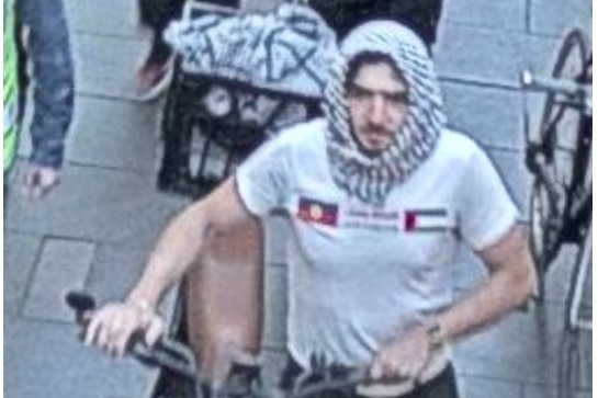 A man wearing a white t-shit and a head scarf wheels a bike through a crowd near the Sydney Opera House