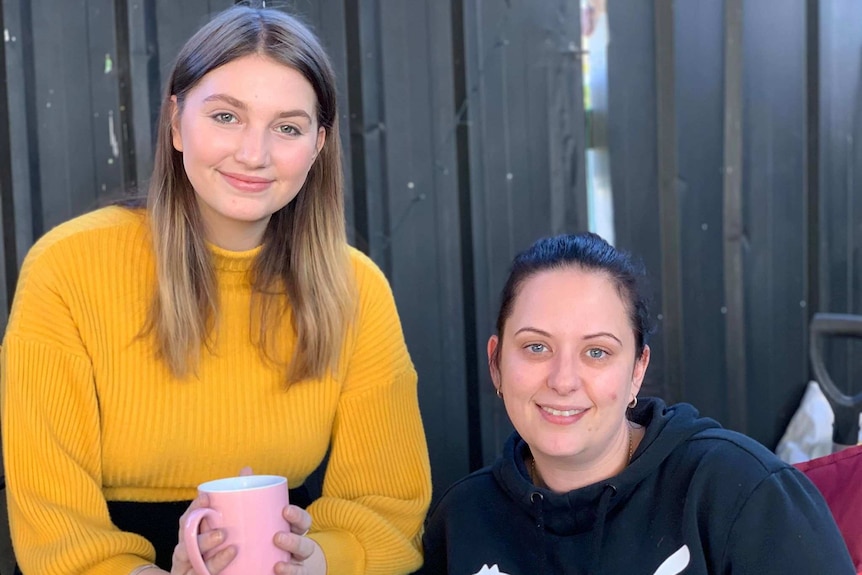 Two women sit together, one holding a mug