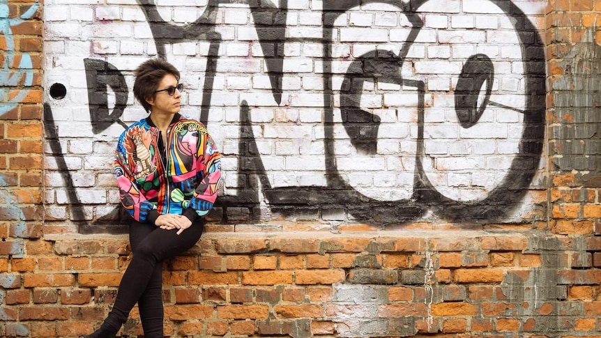 Woman wearing a colourful jacket sitting with a wall behind her with the word Dingo sprayed in graffiti behind her.
