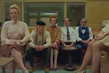 A row of five people - men and women - dressed in 50s office attire sit in a row within a small office of the era.