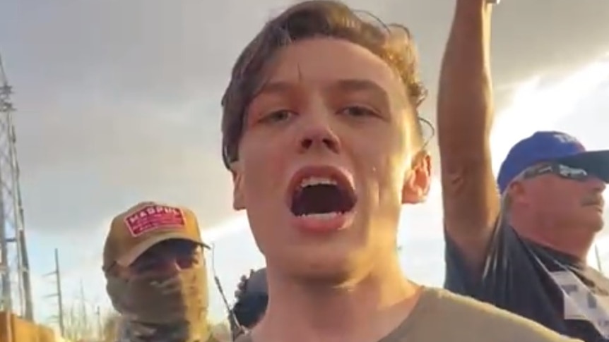 A young man with his mouth open staring at the camera with protesters behind him