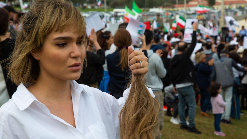 A woman in a white collared shirt holds a lock of her hair at Bondi Beach in protest, with many other protesters