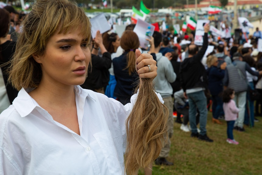 A woman in a white collared shirt holds a lock of her hair at Bondi Beach in protest, with many other protesters